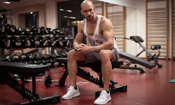 Strong, determined man in white tank top sitting in gym, showcasing fitness and strength.